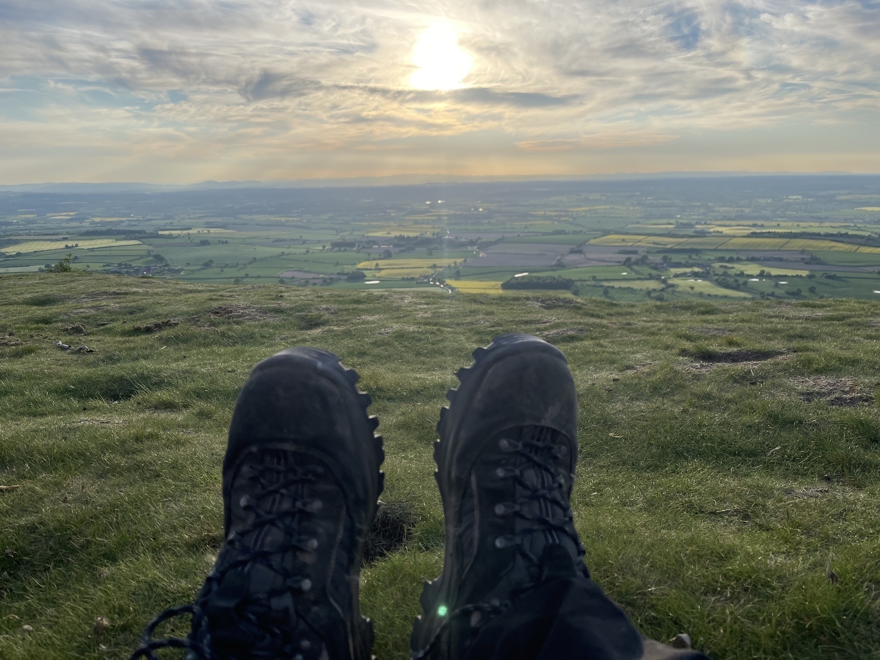 Picture taken of the photographers legs while sitting on the Wrekin, showing his walking boots in the center lower part of the photo, overlooking the patchwork fields of the Shropshire countryside with the sun showing through the clouds at sunset.